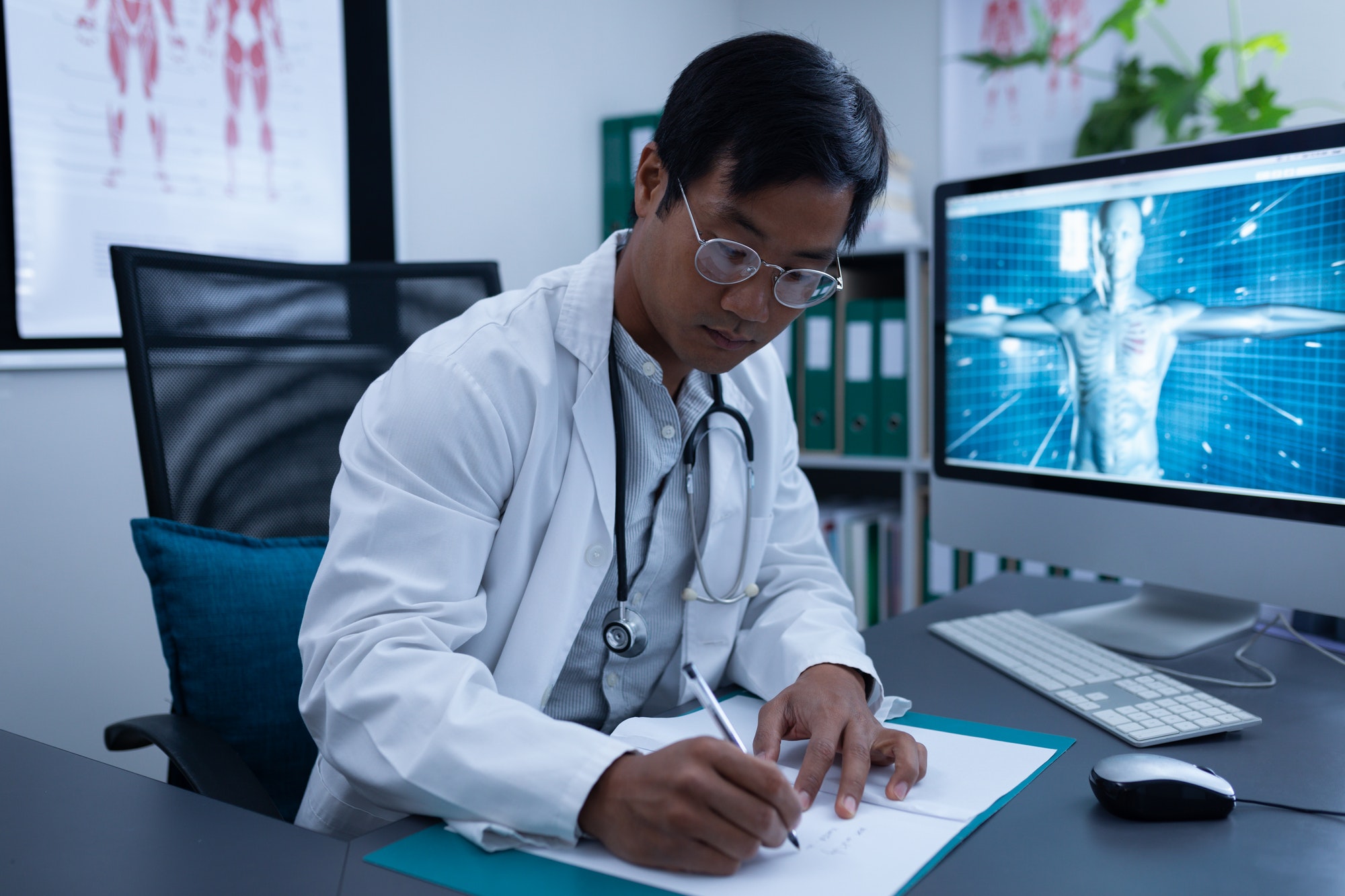 Front view of confident Asian male doctor writing on medical file in clinic at hospital