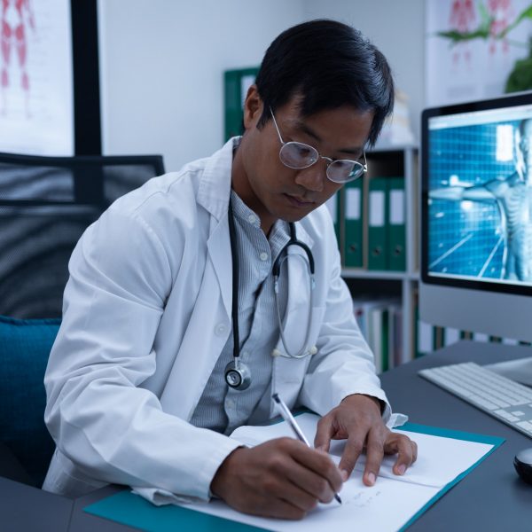 Front view of confident Asian male doctor writing on medical file in clinic at hospital